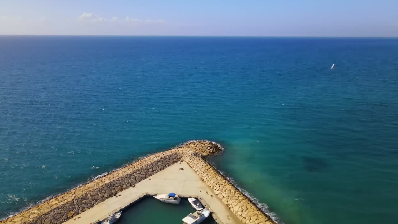 Drone shot showing protected marina enclosed by stone breakwaters, small boats docked inside and single sailboat visible beyond calm blue waters off the coast of Durres
