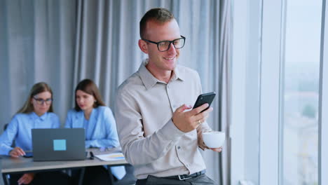 relaxed middle-aged male drinking coffee using phone. happy smiling office employee close up. women colleagues working on laptop at backdrop in blur.