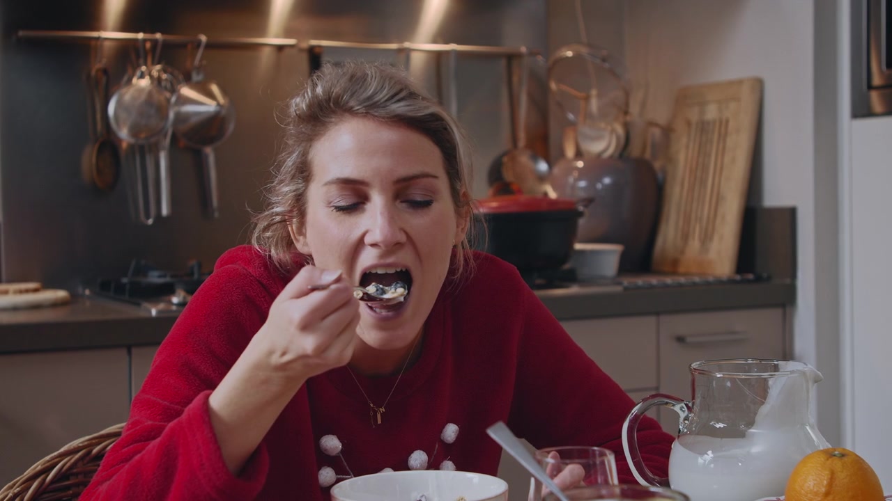 Ung kvinne sitter ved et dekket bord på kjøkkenet sitt, tilbereder og spiser en sunn frokost med cruesli og yoghurt, omgitt av julepynt og festbestikk, og koser seg.