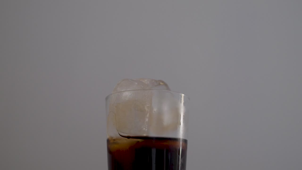 Un bicchiere di cola con cubetti di ghiaccio su uno sfondo neutro.