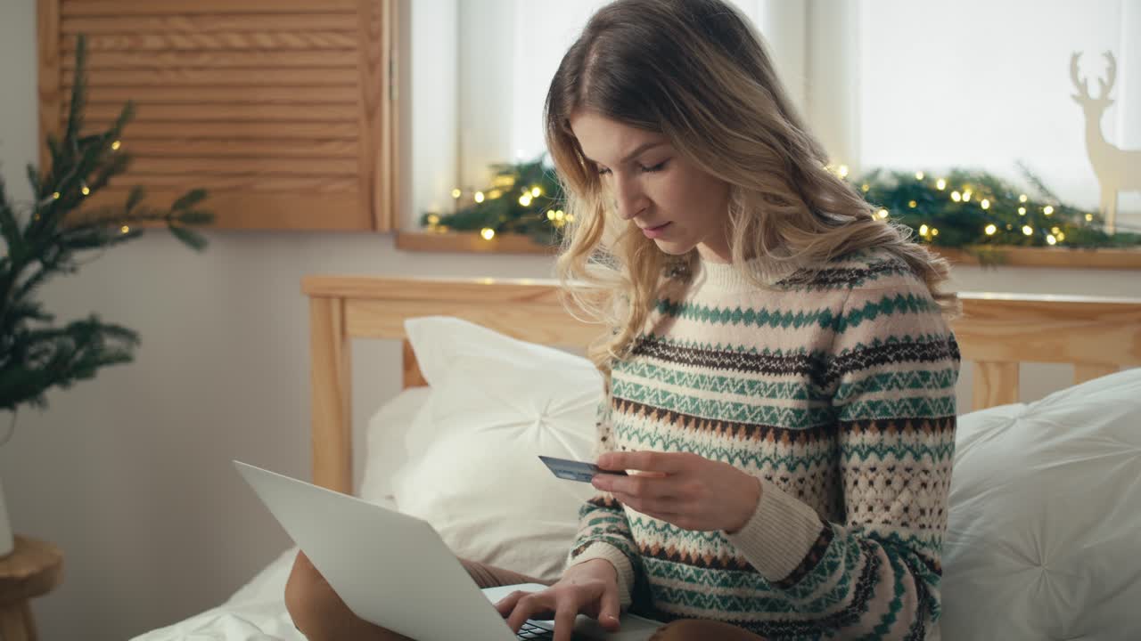 mujer caucásica sentada en la cama y haciendo compras de navidad con el uso de una computadora portátil y tarjeta de crédito.