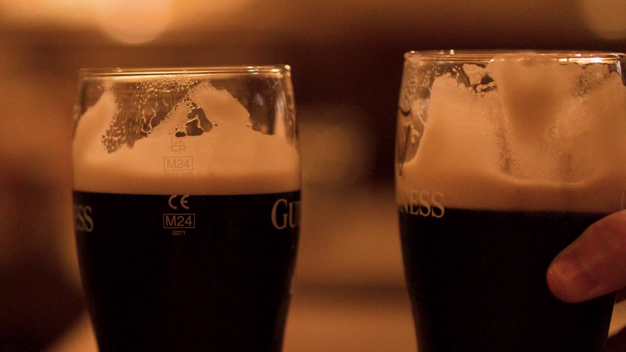 Two dark beers clink together in warm, ambient bar lighting with shallow depth of field