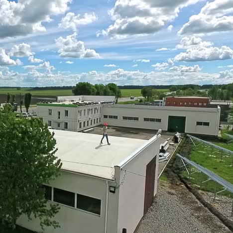 Technician in orange helmet on the roof. Worker goes on a flat roof of industrial building on the background of a construction of a new solar farm. Aerial view.