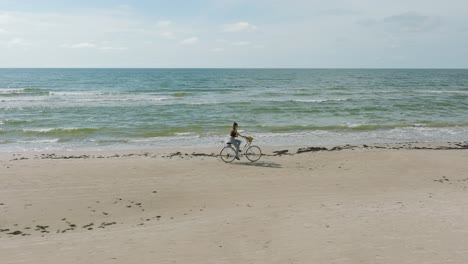 Aerial-view-with-a-young-longhaired-girl-riding-a-bike-on-the-sandy-beach,-sunny-day,-white-sand-beach,-active-lifestyle-concept,-wide-drone-dolly-shot-moving-right