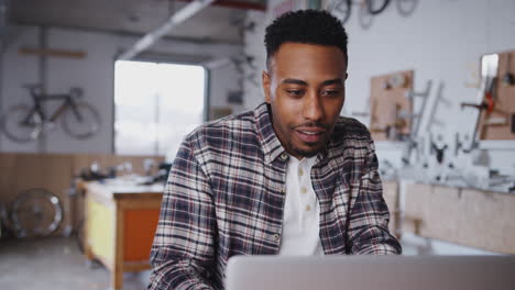 young male business owner in workshop for sustainable bicycles doing accounts on laptop