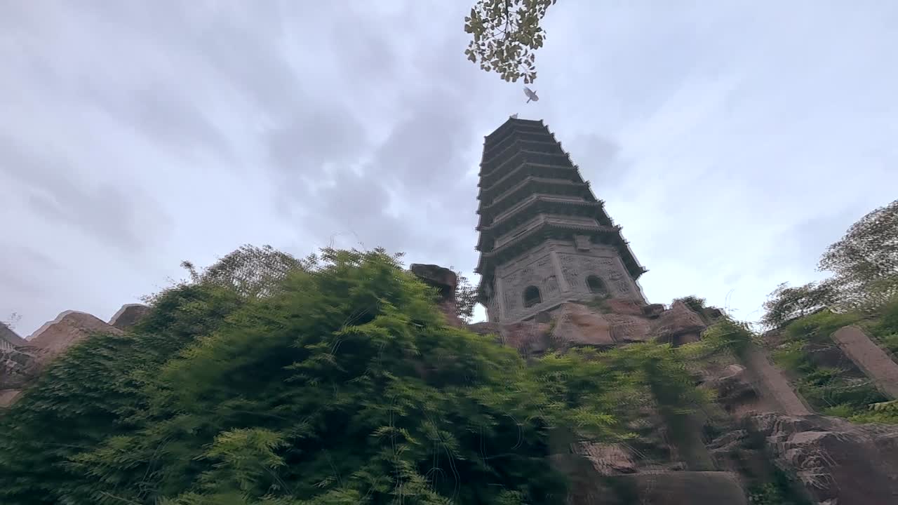 A tall, ancient-looking pagoda is framed by large rocks and lush green foliage, with a cloudy sky in the background