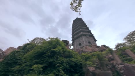 a tall, ancient-looking pagoda is framed by large rocks and lush green foliage, with a cloudy sky in the background