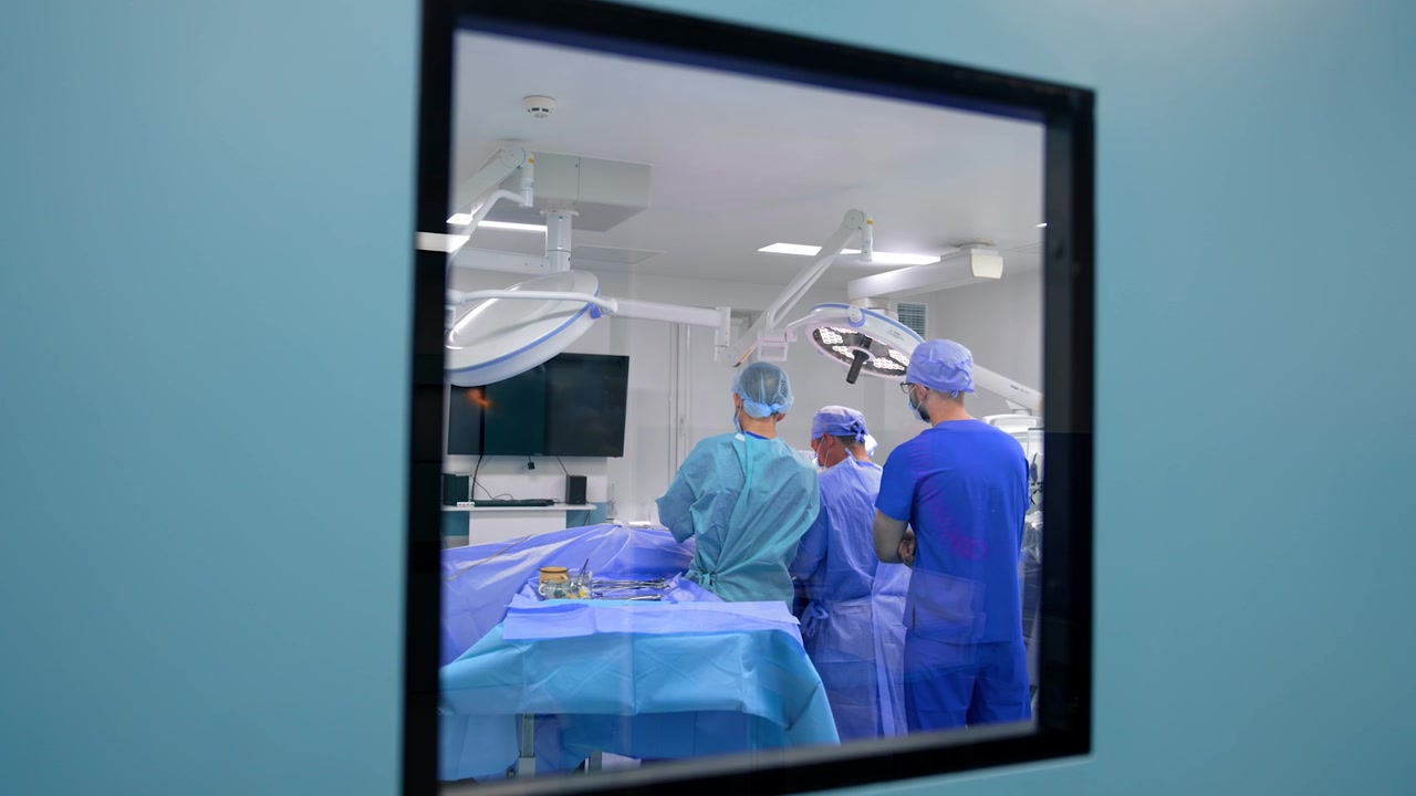 Puerta azul con ventana a una moderna sala de cirugía. Equipo de médicos trabaja en la sala operando a un paciente