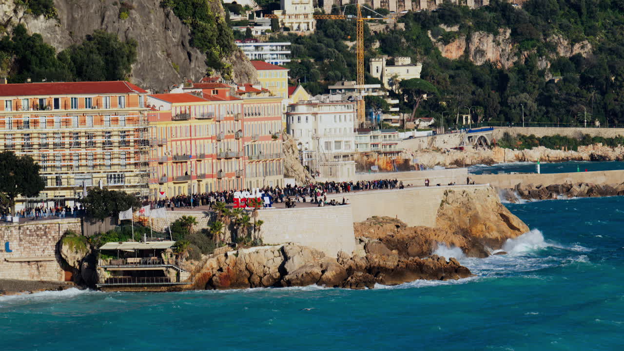Nice, France - March 17, 2025: View of people walking on the coast of the city with water crashing on the rocks and the buildings on the background