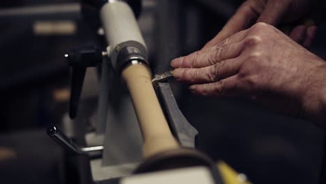 Slow-motion-carpenter-wrinkled-hands-cutting-wooden-knob-out-of-wood-piece-spinning-on-machine-using-chisel,-close-up-shot