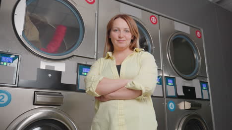 smiling laundress stands before labelled industrial cloth dryers, clothes rolling behind glass, stainless drums and glowing digital controls in modern laundromat, showcasing service