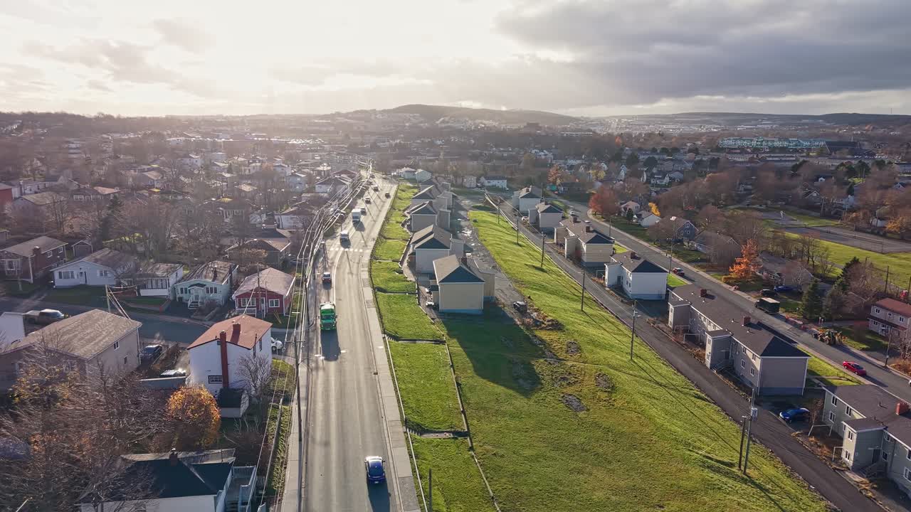 Aerial view of a suburban street lined with neat houses, green lawns and autumn trees, stretching toward distant hills under a partly cloudy sky