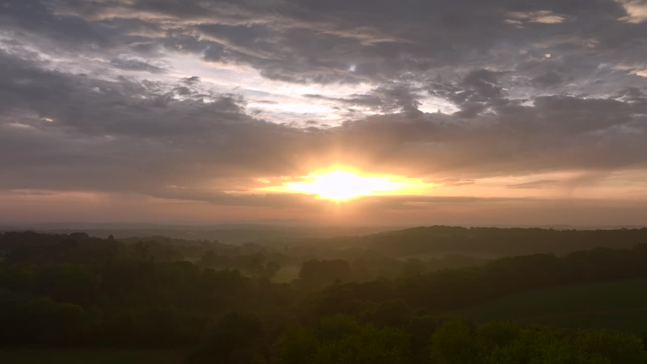 Serene sunrise over rolling patchwork fields and misty hedgerows and woodland. Summer. Cornwall, UK.