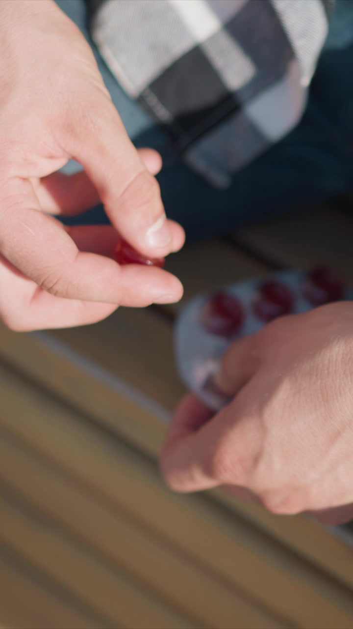 Primo piano di mani che aprono un blister con pillole rosse, rivelando dettagli della texture e illuminazione soffusa. Le mani manipolano la confezione contro uno sfondo di superficie in legno, mostrando un'azione deliberata.