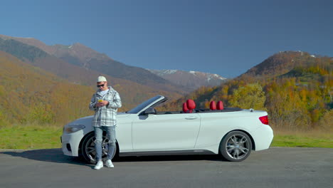 man using phone next to a white convertible car in the mountains during autumn