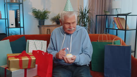 Happy-senior-old-man-celebrating-birthday-party,-makes-wish-blowing-burning-candle-on-small-cupcake