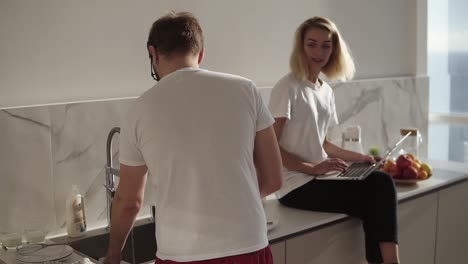 couple spending time together in talk at the kitchen. blonde woman using a laptop, sitting on a kitchen counter while her husband is washing the dishes. morning conversation. slow motion