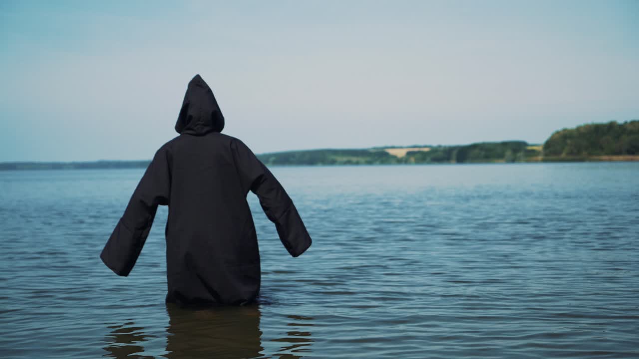 Scary figure in black mantle in the river. Mysticism of Halloween. Halloween costume.