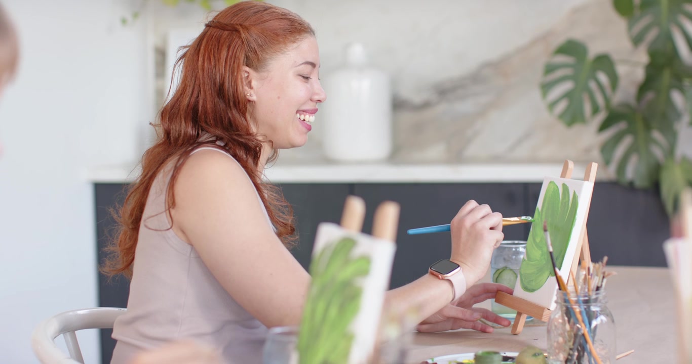 Diverse female friends picking brush dipping in jar painting green leaves on easel while learning