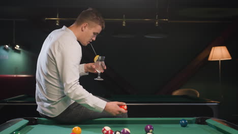 player in white shirt and checkered trousers sits on green pool table, sipping lemon drink while rolling billiard ball. stylish billiards lounge with ambient lighting creates a relaxed atmosphere