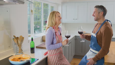 mature couple wearing aprons drinking red wine as they prepare ingredients and cook meal at home together - shot in slow motion