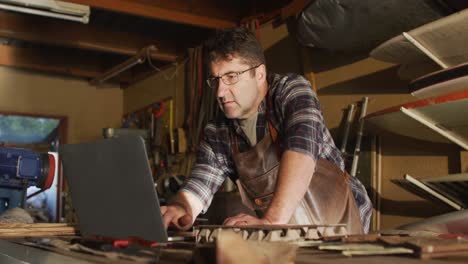 Focused-caucasian-male-knife-maker-in-workshop-using-laptop