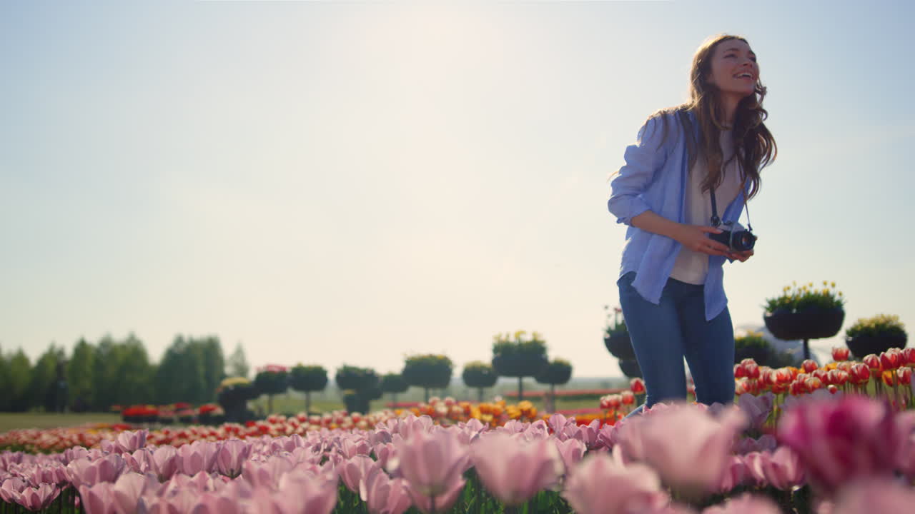 mujer feliz tocando flores a pie con la cámara en el campo de tulipanes en un día soleado.