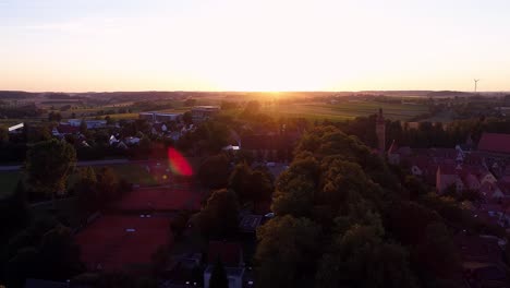 Luftdrohne-Senkt-Sich-Sanft-Gegen-Den-Sonnenuntergang,-Wunderschönes-Panorama-Der-Altstadt,-Fußballplatz,-Bäume,-Park,-Grüne-Wiesen-Und-Felder,-Linseneffekt