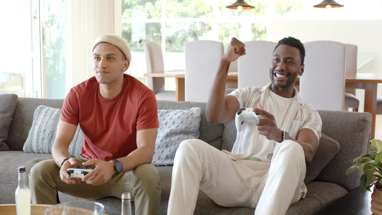 Hispanic and African American friends grabbing controllers, playing on sofa with chips cheering win