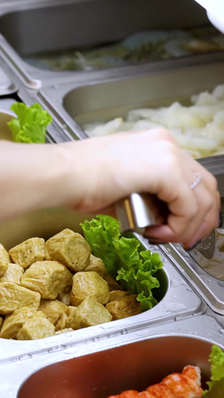 Una mano seleziona ingredienti assortiti per hotpot da vassoi buffet in acciaio inossidabile sotto la luminosa illuminazione interna di un ristorante.
