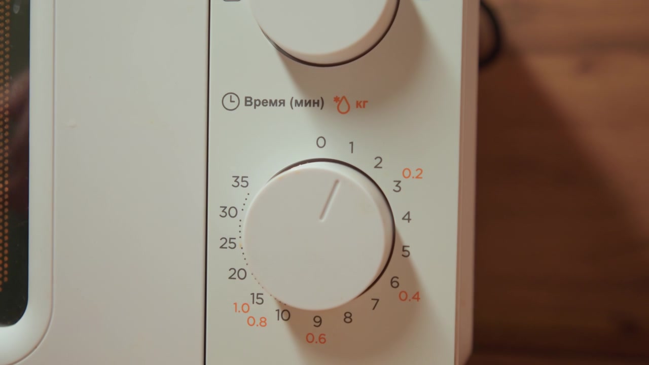 Primo piano di una mano che gira una manopola rotante sul pannello di controllo di un microonde bianco per regolare il timer o l'impostazione della funzione; tre quadranti circolari impilati con numeri e simboli stampati visibili, piano di lavoro in legno.