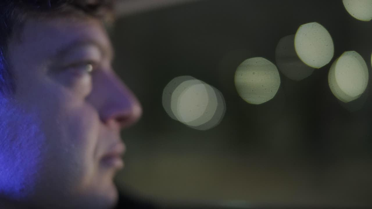 Slow motion close up of young handsome businessman looking outside during traveling in a modern car with a driver in center of the city by the night. City lights
