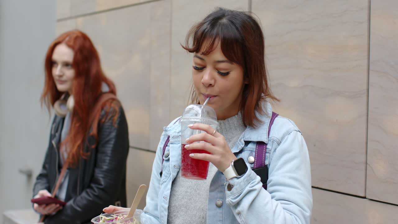 Notando una bevanda rossa ghiacciata, una donna la assaggia con la cannuccia tra un boccone e l'altro di insalata su una passeggiata urbana.
