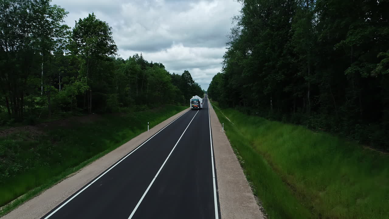 흐린 날 시골을 따라 도로를 따라 운전하는 동안 목조 주택을 운반하는 트럭 위로 공중 드론이 뒤로 이동하는 샷
