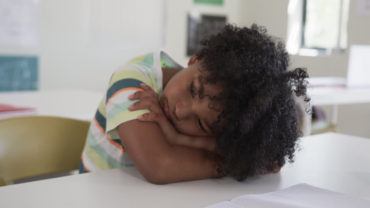 Bambino afroamericano che si riposa con le braccia sul tavolo di una classe, mentre guarda dei fogli e una matita dopo aver studiato