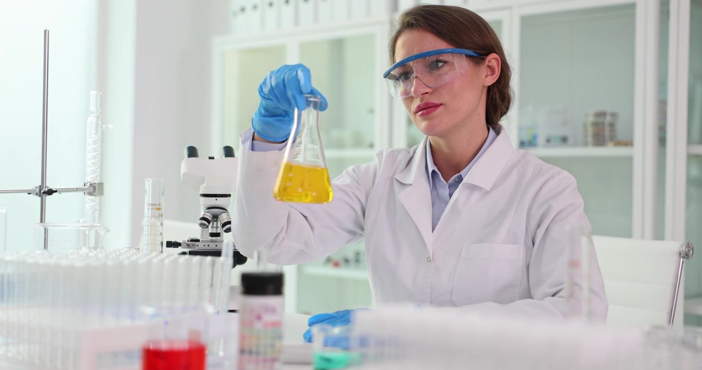 Focused female scientist shakes flask with yellow liquid in lab