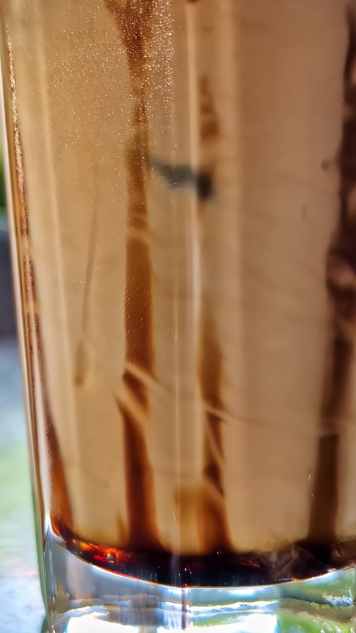 Caffè e panna si mescolano in una tazza di vetro, creando motivi fluidi e scorrevoli con ricche tonalità marroni in una morbida luce naturale. Video verticale, ideale per i social media.