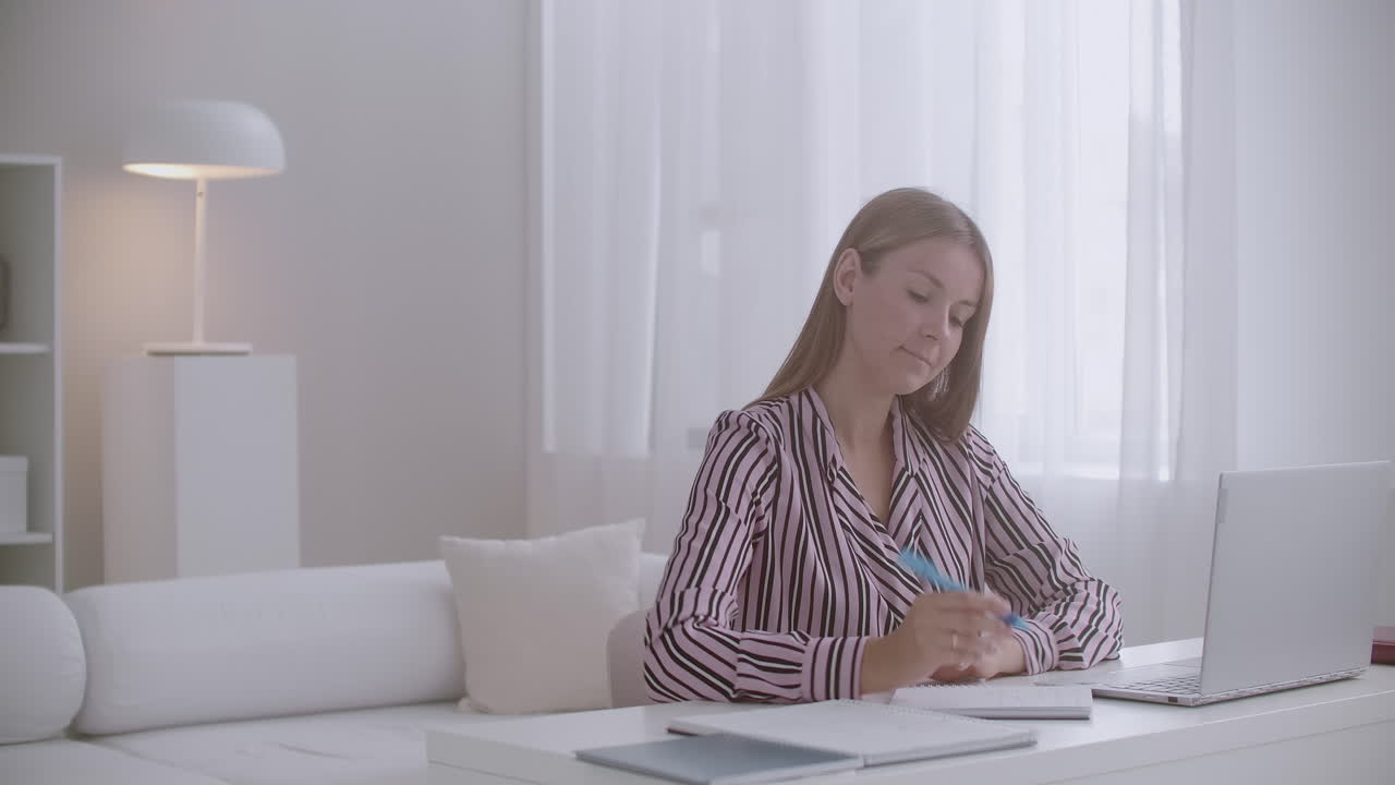 예쁜 젊은 학생 여성은 집에서 온라인으로 공부하고 테이블에 앉아 노트북에서 웹 세미나를 보고 메모를 쓰고 있습니다.