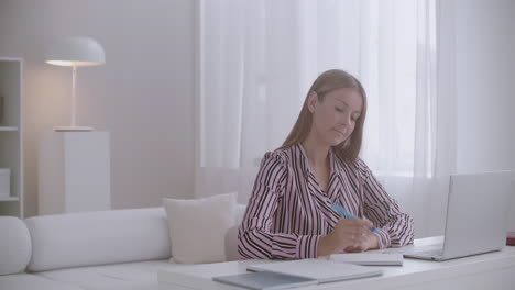 young pretty student woman is learning online at home sitting at table watching webinar at laptop and writing notes