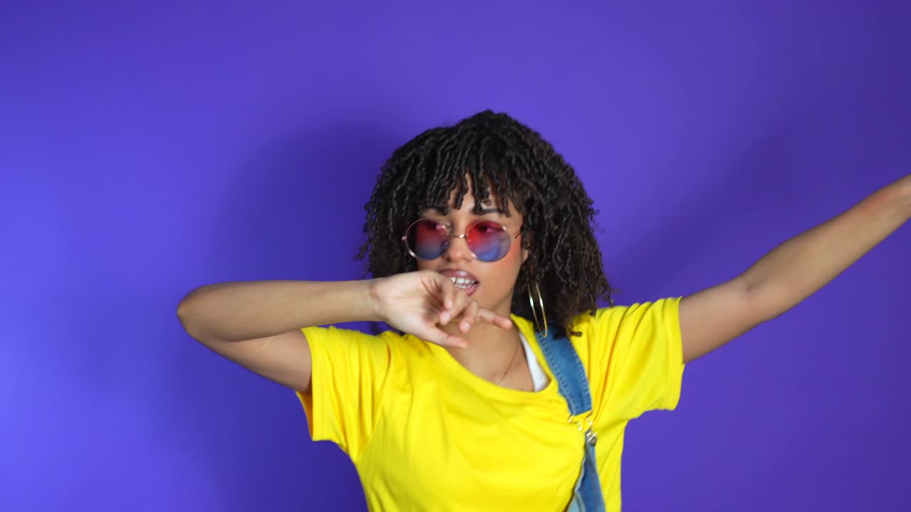 Kvinde i gul trøje og denim overalls, der danser foran en lilla baggrund