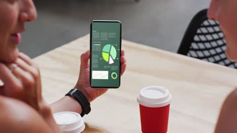 Caucasian-man-and-woman-sitting-at-table,-using-smartphone-with-statistics-on-screen