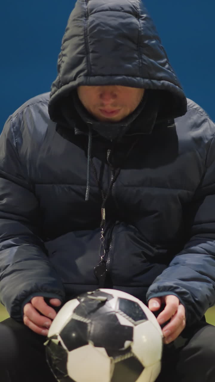 un hombre se sienta con la cabeza hacia abajo, sosteniendo una pelota de fútbol desgastada entre sus piernas, el fondo presenta una vista borrosa de un estadio y un poste de gol