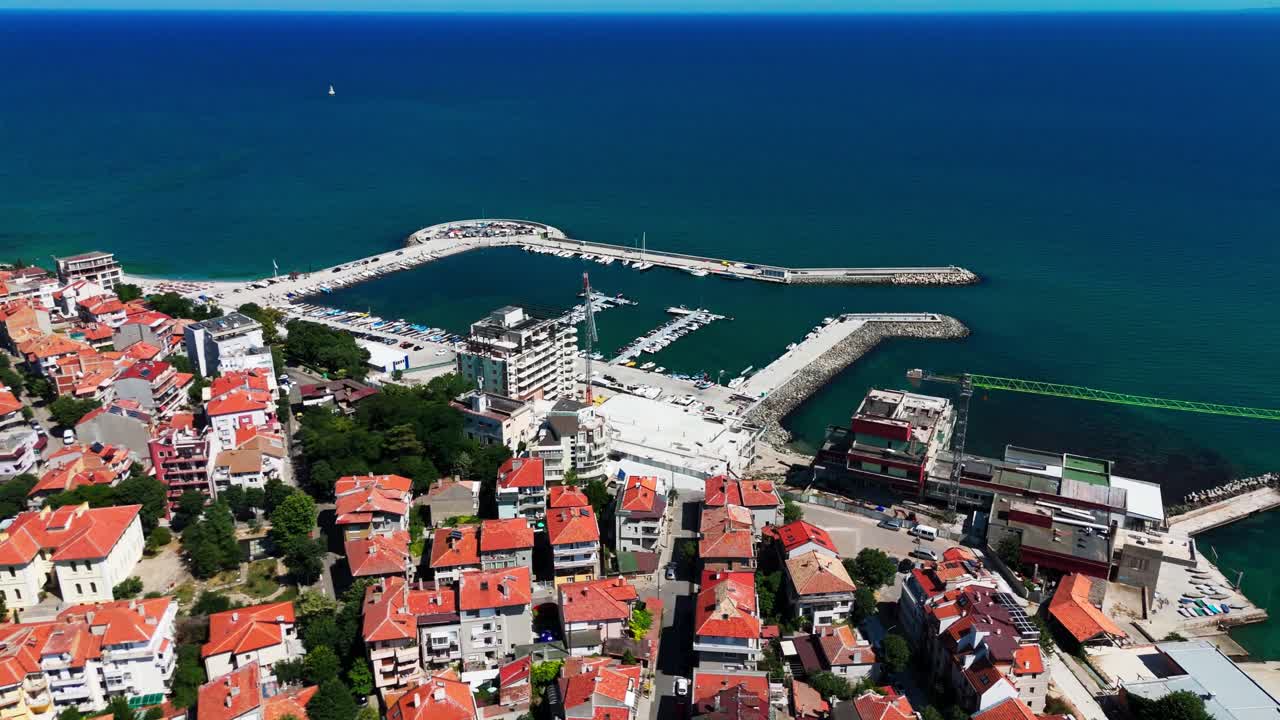 Dron se blíží k jednomu z hlavních přístavů Pomorie s loděmi, střechami a otevřeným Černým mořem v pozadí.