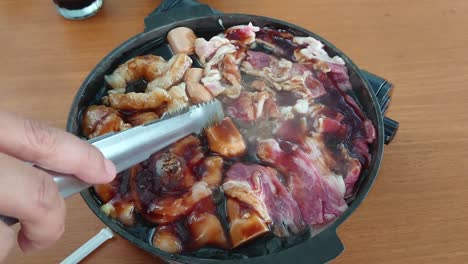 tasty delicious japanese style meats cooking, grilling, sizzling, and frying in interior black pan with teriyaki sauce in kitchen, rotated with caucasian fingers holding silver tongs, overhead static