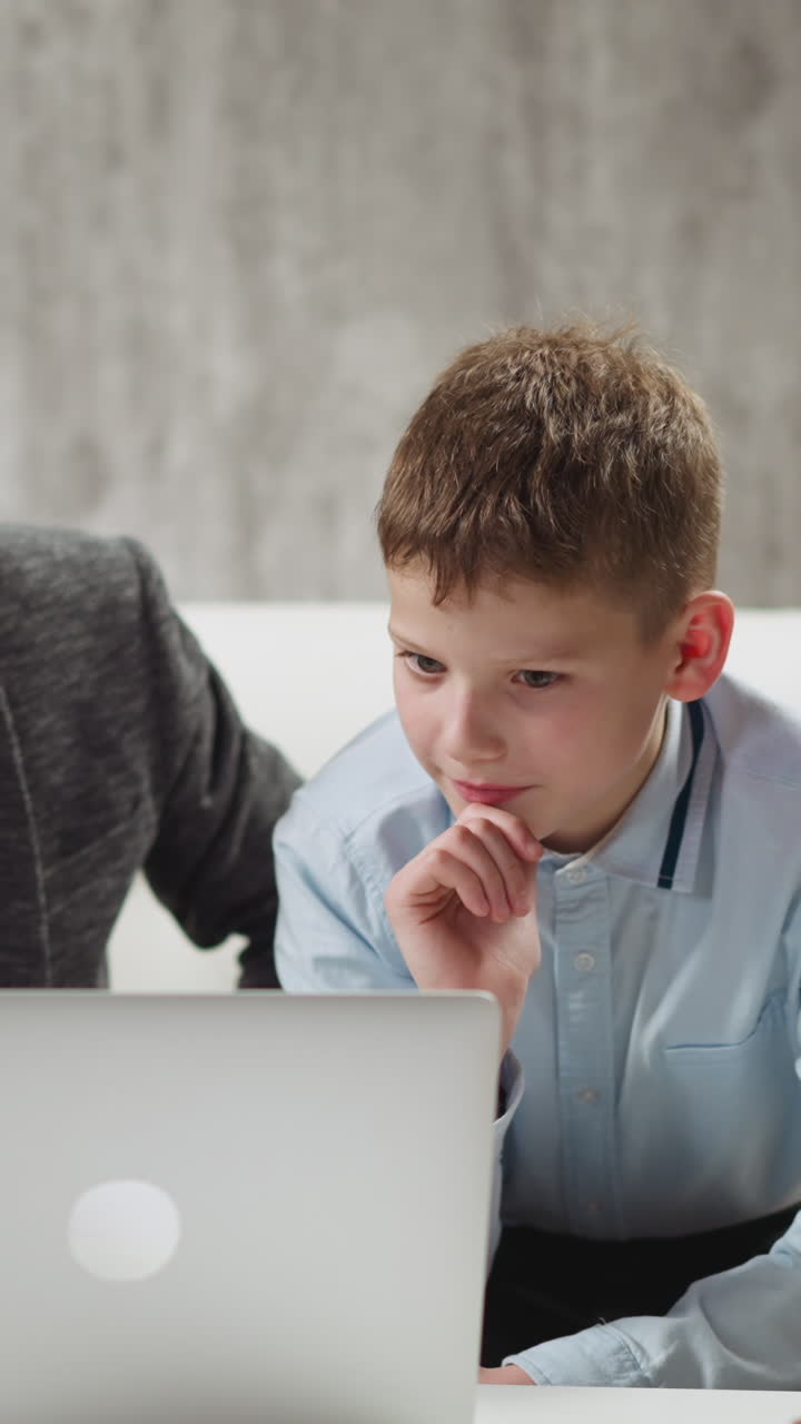 Concentrated boy student watches educative video via laptop and declines careful English teacher help at home private lesson slow motion