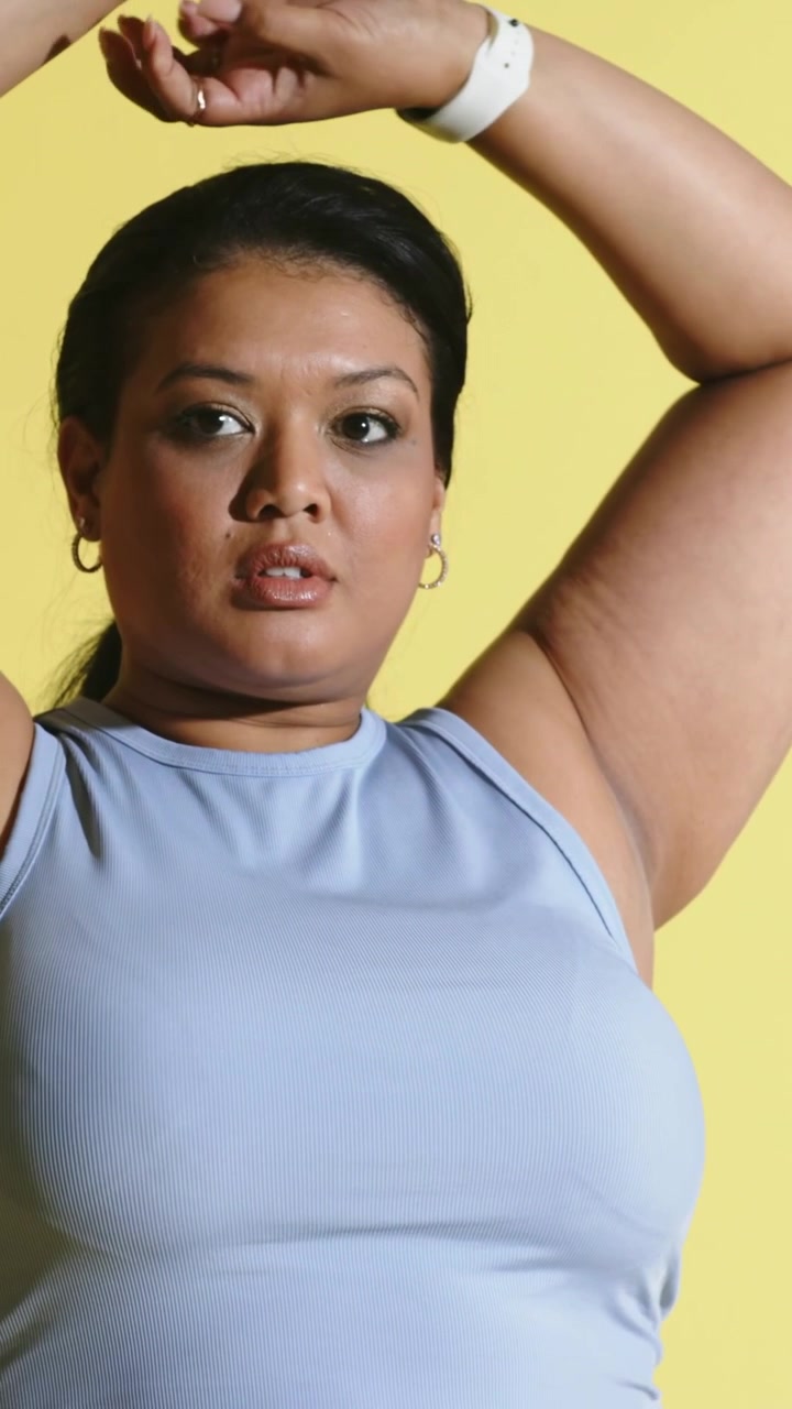 Video verticale: la telecamera si allontana da una donna con crop top blu che alza le braccia su un set giallo con un orologio.