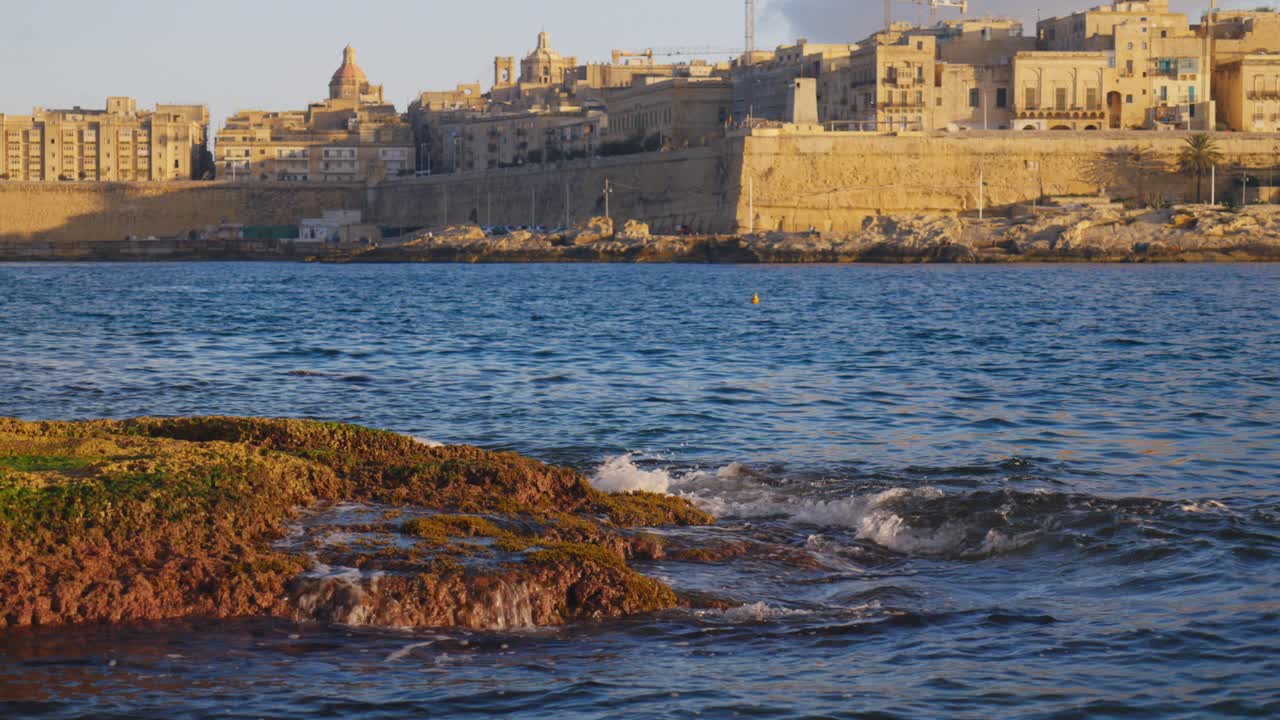 Onde del mare che si infrangono sulle rocce costiere con lo skyline di La Valletta sullo sfondo all'ora d'oro, Malta.