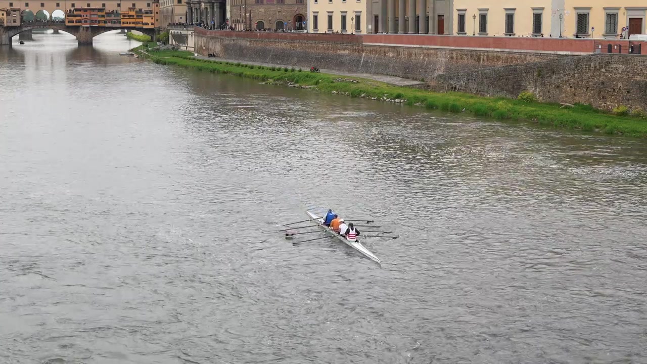 Wide shot of a rowing crew gliding down the Arno River in Florence, Italy, with the iconic Ponte Vecchio and classic architecture lining the water under soft daylight.