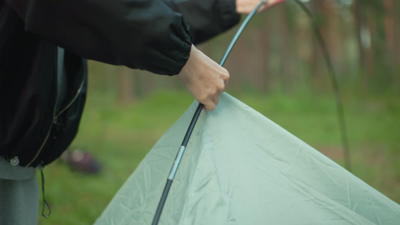 close up of woman in black jacket struggling to remove tent hook from flexible tent pole while another person hand assists partially visible in forest environment surrounded by green grass and trees