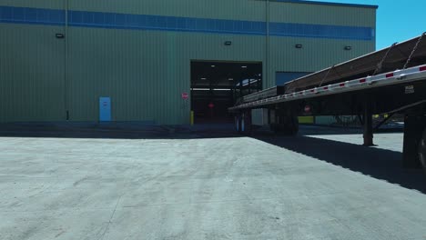 low angle dolly follows semi truck with steel beams reversing into warehouse at midday